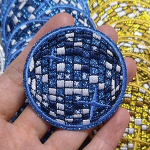 Op de afbeelding: Een ronde, blauw-witte glitter strijkpatch met een geruit patroon en stervormige ontwerpen. De patch is rond en heeft een blauwe rand.