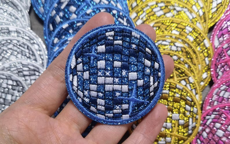 Op de afbeelding: Een ronde, blauw-witte glitter strijkpatch met een geruit patroon en stervormige ontwerpen. De patch is rond en heeft een blauwe rand.
