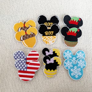 May include: Set of ten embroidered iron-on patches featuring various designs, including Mickey Mouse, acorns, snowflakes, and American flags. The patches are colourful and perfect for adding a personal touch to clothing, bags, or other fabric items.