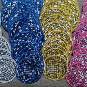 Op de afbeelding: Een verzameling ronde, glimmende, stoffen patches in verschillende kleuren, waaronder zilver, blauw, goud en roze. Elke patch heeft een gespiegelde, moza&iuml;ekachtige vormgeving.