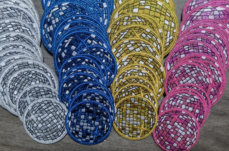 Op de afbeelding: Een verzameling ronde, glimmende, stoffen patches in verschillende kleuren, waaronder zilver, blauw, goud en roze. Elke patch heeft een gespiegelde, moza&iuml;ekachtige vormgeving.
