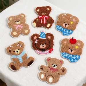 Puede incluir: Una colección de siete parches bordados de ositos de peluche con tela marrón texturizada. Cada oso tiene detalles únicos que incluyen lazos, bufandas, gorros de fiesta rosas, rojos o azules y pequeños adornos como estrellas o clips.