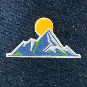 Könnte beinhalten: Ein gestickter Aufnäher, der eine blaue und weiße Bergkette mit einer gelben Sonne darüber zeigt. Die Bergkette hat grüne Akzente.