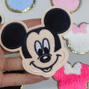 May include: A beige felt Mickey Mouse character patch with black ears and eyes. The patch has a stitched smile and a pink tongue.