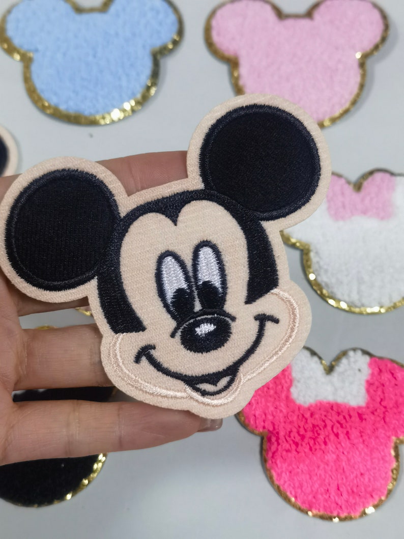 May include: A beige felt Mickey Mouse character patch with black ears and eyes. The patch has a stitched smile and a pink tongue.