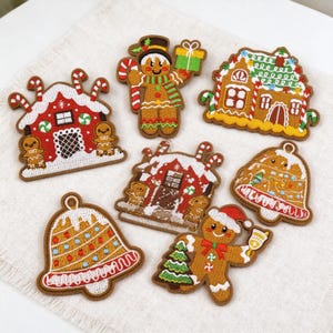 Op de afbeelding: Een verzameling van zeven geborduurde patches met kerstthema's, waaronder twee peperkoekhuisjes met rode en bruine details, twee peperkoekmannetjes en twee klokvormige ornamenten met kleurrijke accenten op een witte stoffen achtergrond.