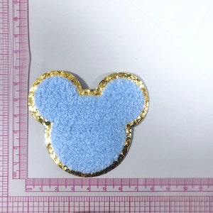 May include: A light blue fuzzy fabric patch shaped like Mickey Mouse ears with a gold glitter outline. The patch is about 5 cm wide.