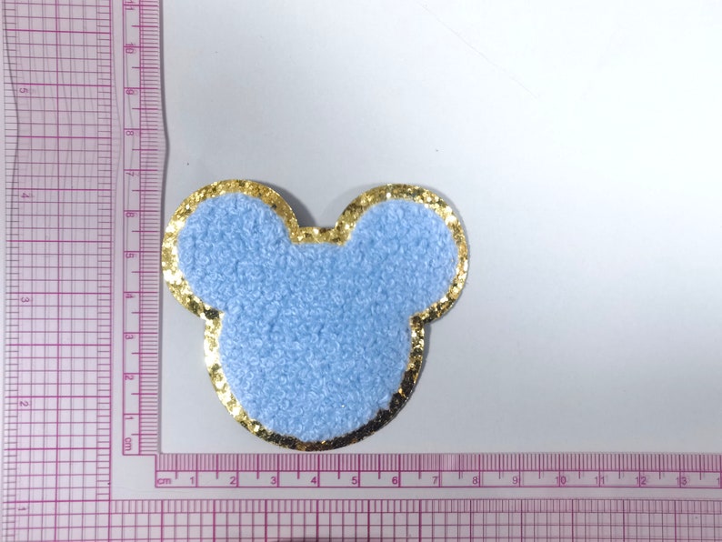 May include: A light blue fuzzy fabric patch shaped like Mickey Mouse ears with a gold glitter outline. The patch is about 5 cm wide.