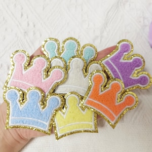 May include: A set of six colourful felt crown patches with gold glitter trim. The crowns are in various colours, including pink, blue, yellow, orange, purple and white.