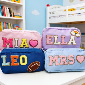 May include: Four rectangular nylon pouches in pink, lavender, navy blue, and light blue. Each pouch features large, colorful, glitter-bordered varsity-style letter patches spelling out the names MIA, ELLA, LEO, and MRS, with additional heart, rainbow, and football patches.
