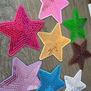 May include: A collection of eight star-shaped patches covered in shiny sequins in various colours including hot pink, light pink, green, gold, brown, lavender, blue, and silver. Each star has a distinct border and a reflective, textured surface.