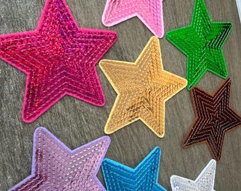 May include: A collection of eight star-shaped patches covered in shiny sequins in various colors including hot pink, light pink, green, gold, brown, lavender, blue, and silver. Each star has a distinct border and a reflective, textured surface.