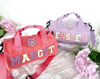 Puede incluir: Dos bolsas de deporte rosa y morado con nombres personalizados, "Margot" y "Ensley", en letras de colores. Cada bolsa tiene un pequeño parche bordado, una flor en la bolsa rosa y una estrella en la bolsa morada.