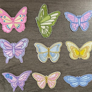 May include: A collection of eight embroidered butterfly patches in various colours including pink, green, blue, purple, and yellow. Each patch features detailed stitching and distinct wing patterns, arranged in a grid on a dark wood surface.