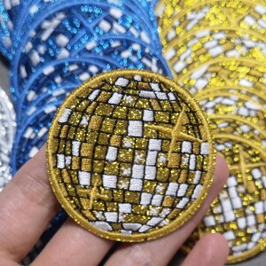 Op de afbeelding: Een close-up van een gouden glitter strijk-op patch in de vorm van een discobal. De patch is omringd door andere patches in verschillende kleuren.