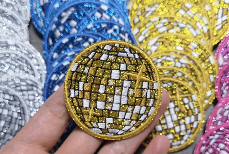 Op de afbeelding: Een close-up van een gouden glitter strijk-op patch in de vorm van een discobal. De patch is omringd door andere patches in verschillende kleuren.