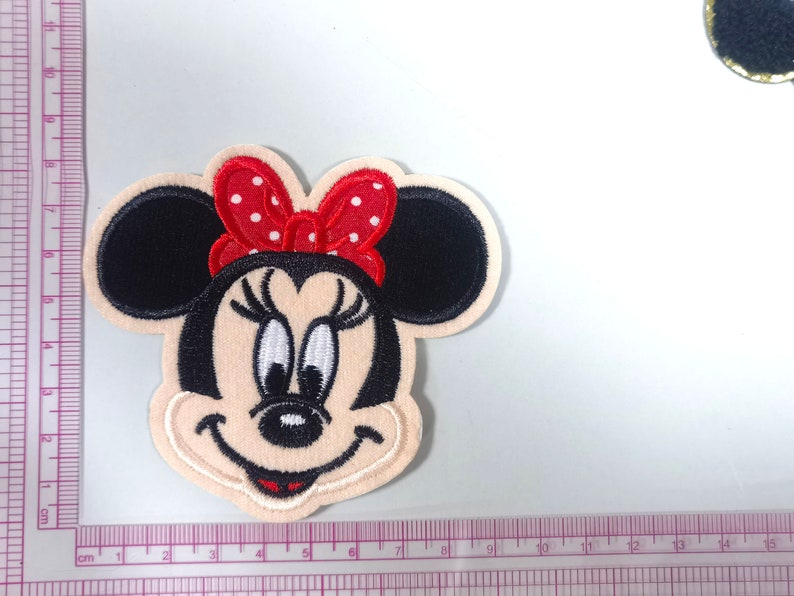 May include: Embroidered iron-on patch featuring a cartoon image of Minnie Mouse with a red bow with white polka dots. The patch is approximately 5 cm in diameter.