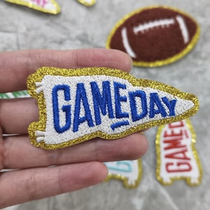 Puede incluir: Un banderín de fieltro blanco con ribete de purpurina dorada y texto bordado en azul que dice "GAME DAY".
