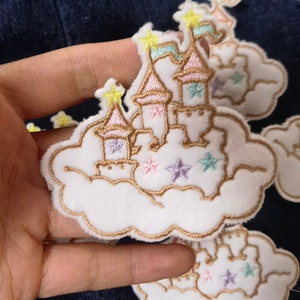 Pode incluir: Um remendo de feltro branco com um contorno bordado a ouro de um castelo numa nuvem. O castelo tem detalhes em rosa, azul e amarelo e é decorado com estrelas.