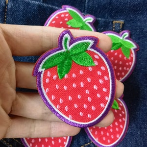 Puede incluir: Un conjunto de cinco parches bordados para planchar que representan fresas rojas con hojas verdes y semillas blancas. Las fresas tienen un contorno morado y están rodeadas de un contorno blanco.