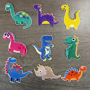 May include: A set of nine embroidered dinosaur patches in various colors including green, purple, yellow, pink, blue, and orange. Each patch features a cartoon-style dinosaur with distinct shapes, including long-necked, spiked, and horned varieties.