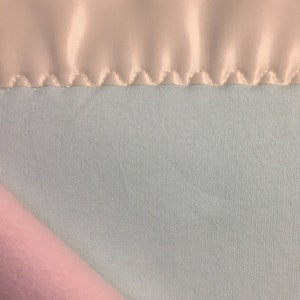 May include: A soft, light blue blanket with a white satin trim. The blanket is folded in half, showing the soft, plush texture of the fabric.