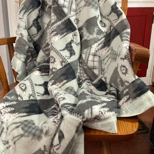 May include: A soft, plush throw blanket with a patchwork design featuring a light green background and gray and white deer silhouettes. The blanket has a light green satin trim.