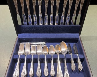 May include: A set of ornate silver-colored flatware, including forks, spoons, and knives, arranged in a dark wooden box with a blue velvet lining. The silverware features detailed floral patterns.