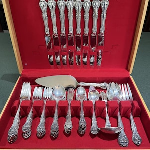 May include: A vintage silverware set in a wooden case with a red velvet interior. The set includes knives, forks, spoons, and serving utensils. The silverware has ornate, floral-patterned handles and a shiny, reflective surface. The case has a label that reads "SHEIPCO".