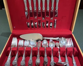 May include: A vintage silverware set in a wooden case with a red velvet interior. The set includes knives, forks, spoons, and serving utensils. The silverware has ornate, floral-patterned handles and a shiny, reflective surface. The case has a label that reads "SHEIPCO".