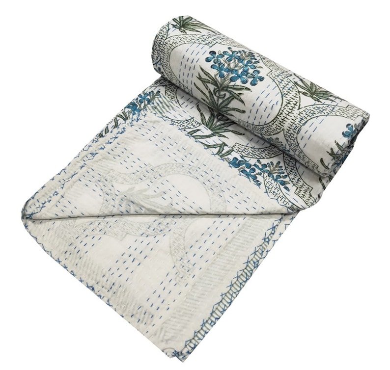 May include: White kantha quilt with a blue and green floral pattern. The quilt is folded and rolled up, showing the stitching detail.