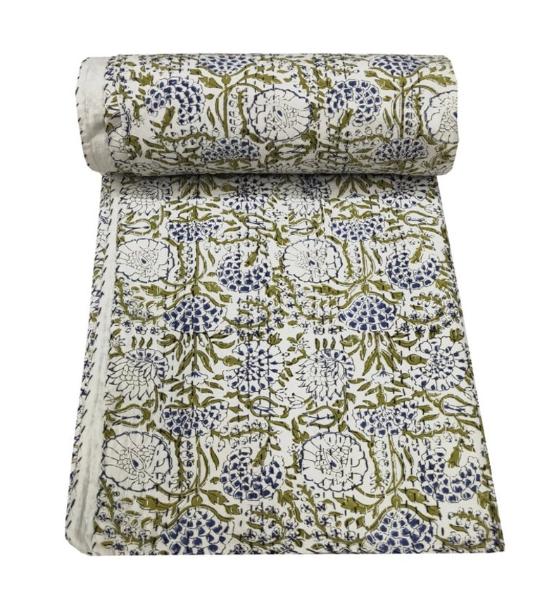 May include: A white cotton quilt with a floral pattern in shades of green and blue. The quilt is rolled up and has a fringed edge.