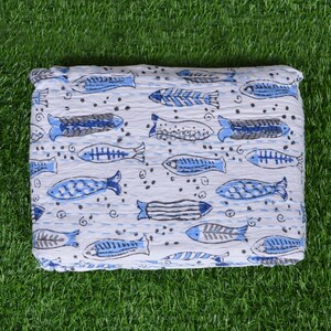 May include: White cotton quilt with a blue and gray fish pattern. The quilt is folded and lying on a green grassy surface.