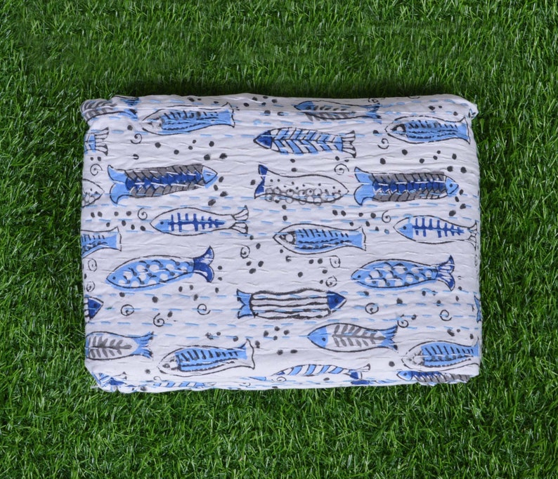 May include: White cotton quilt with a blue and gray fish pattern. The quilt is folded and lying on a green grassy surface.