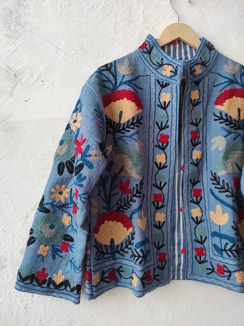 Peut inclure: Veste bleu clair avec broderies florales d&eacute;taill&eacute;es en rouge, jaune et vert. La veste a un col mandarin et une doublure ray&eacute;e. La broderie pr&eacute;sente des fleurs et des feuilles stylis&eacute;es, cr&eacute;ant un design color&eacute;.