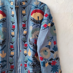 Peut inclure: Veste bleu clair orn&eacute;e de broderies florales d&eacute;taill&eacute;es en rouge, jaune, vert et bleu. La veste a un col mandarin et une doublure ray&eacute;e. La broderie couvre le devant, les manches et le col, avec des motifs floraux.
