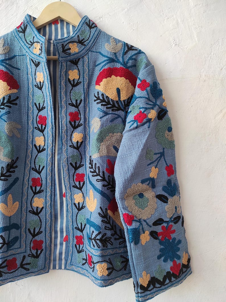 Peut inclure: Veste bleu clair orn&eacute;e de broderies florales d&eacute;taill&eacute;es en rouge, jaune, vert et bleu. La veste a un col mandarin et une doublure ray&eacute;e. La broderie couvre le devant, les manches et le col, avec des motifs floraux.