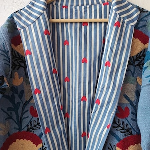 Peut inclure: Veste bleue brod&eacute;e avec un int&eacute;rieur ray&eacute; orn&eacute; de c&oelig;urs rouges. La veste pr&eacute;sente des broderies florales et de feuilles rouges, jaunes et vertes. La veste est suspendue &agrave; un cintre en bois.