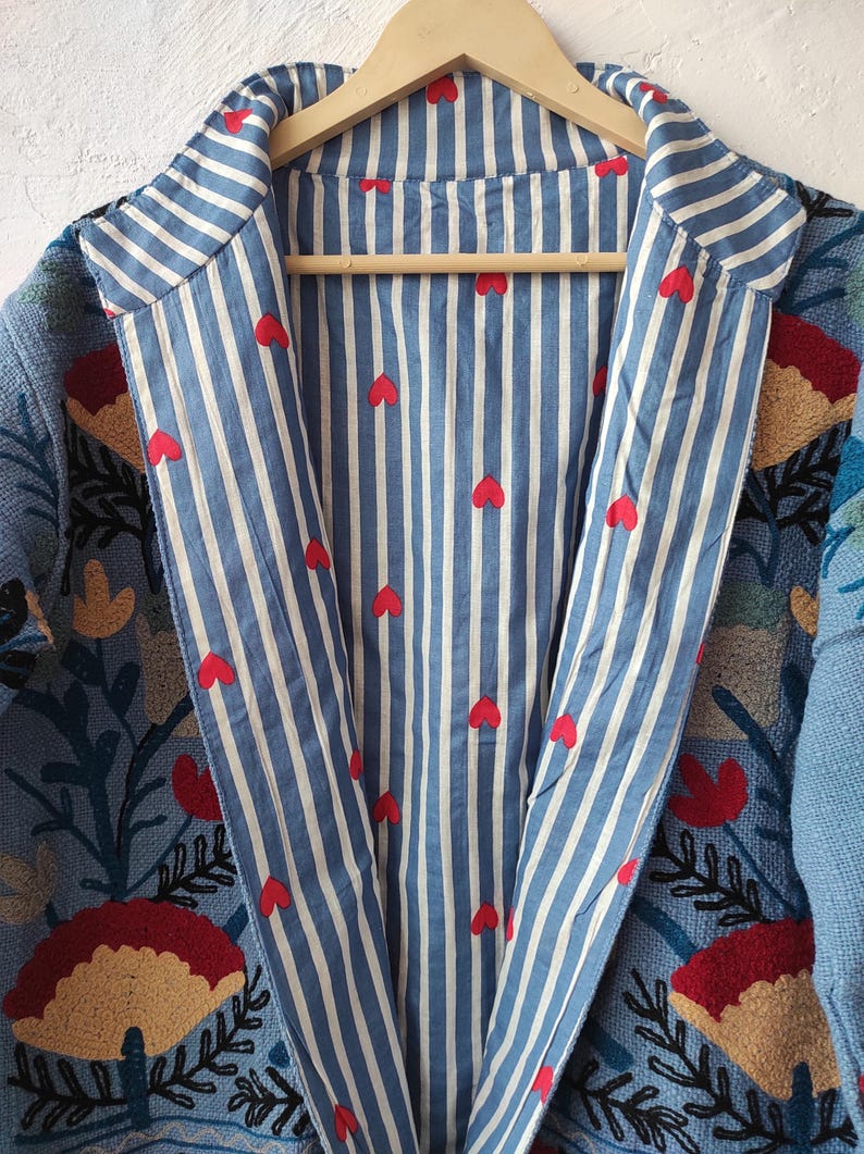 Peut inclure: Veste bleue brod&eacute;e avec un int&eacute;rieur ray&eacute; orn&eacute; de c&oelig;urs rouges. La veste pr&eacute;sente des broderies florales et de feuilles rouges, jaunes et vertes. La veste est suspendue &agrave; un cintre en bois.