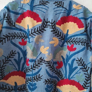 Peut inclure: Veste bleu clair avec broderie florale d&eacute;taill&eacute;e. Le motif pr&eacute;sente des fleurs rouges, beiges et jaunes, ainsi que des feuilles et des tiges vertes et noires. Gros plan sur la couture complexe.