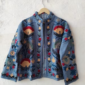 Peut inclure: Veste bleu clair avec broderies florales complexes rouges, jaunes et vertes. La veste a un col mandarin et une doublure ray&eacute;e. Elle est suspendue &agrave; un cintre en bois contre un mur blanc.