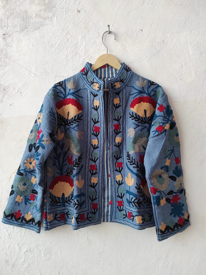 Peut inclure: Veste bleu clair avec broderies florales complexes rouges, jaunes et vertes. La veste a un col mandarin et une doublure ray&eacute;e. Elle est suspendue &agrave; un cintre en bois contre un mur blanc.