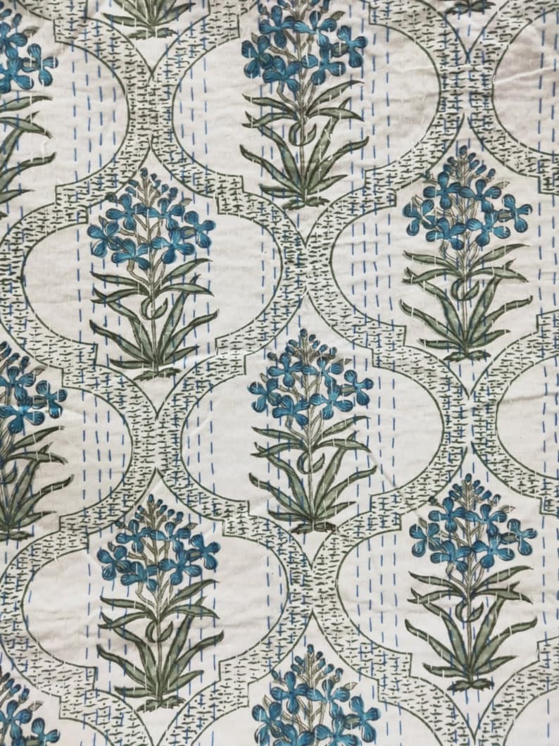 May include: A white fabric with a repeating pattern of blue flowers and green leaves. The flowers are arranged in a symmetrical pattern within a repeating geometric design.