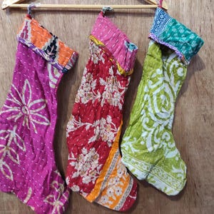 Pode incluir: Três meias de Natal coloridas penduradas em um cabide de madeira. As meias são feitas de tecido estampado em tons de rosa, vermelho e verde. Cada uma tem um design floral único e um punho contrastante.