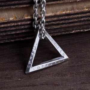 Op de afbeelding: Een zilveren driehoekige hanger aan een ketting. De hanger heeft gegraveerde markeringen langs de randen. De ketting is ook zilverkleurig. De achtergrond is een donker houten oppervlak.