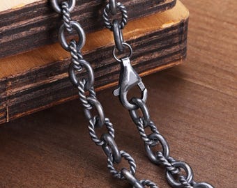 May include: Close-up of a silver-toned chain necklace with a textured, twisted rope design. The chain is connected with oval links and a lobster clasp. The necklace rests on a wooden surface.