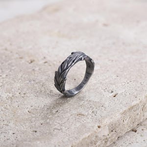 May include: A dark oxidized silver ring featuring a textured leaf or feather pattern along the outer band. The interior of the band is engraved with the text 925 and Psalm 144:1. The ring has a weathered, metallic finish with visible dark detailing.