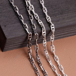 May include: A silver-toned chain necklace featuring a series of elongated, oval-shaped links, each reinforced with a central horizontal bar. The metallic links are interconnected to form a continuous, flexible strand with a polished, reflective finish.