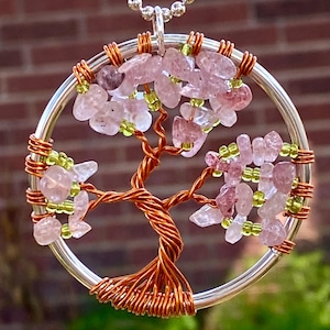May include: A handcrafted pendant necklace featuring a wire-wrapped tree design. The tree has a copper-colored trunk and branches, with pink and green gemstone leaves. The pendant is suspended from a silver-colored chain.