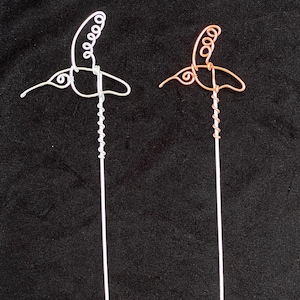 May include: Two wire hummingbird sculptures, one silver and one copper, with a curled tail and a long, thin wire stem.